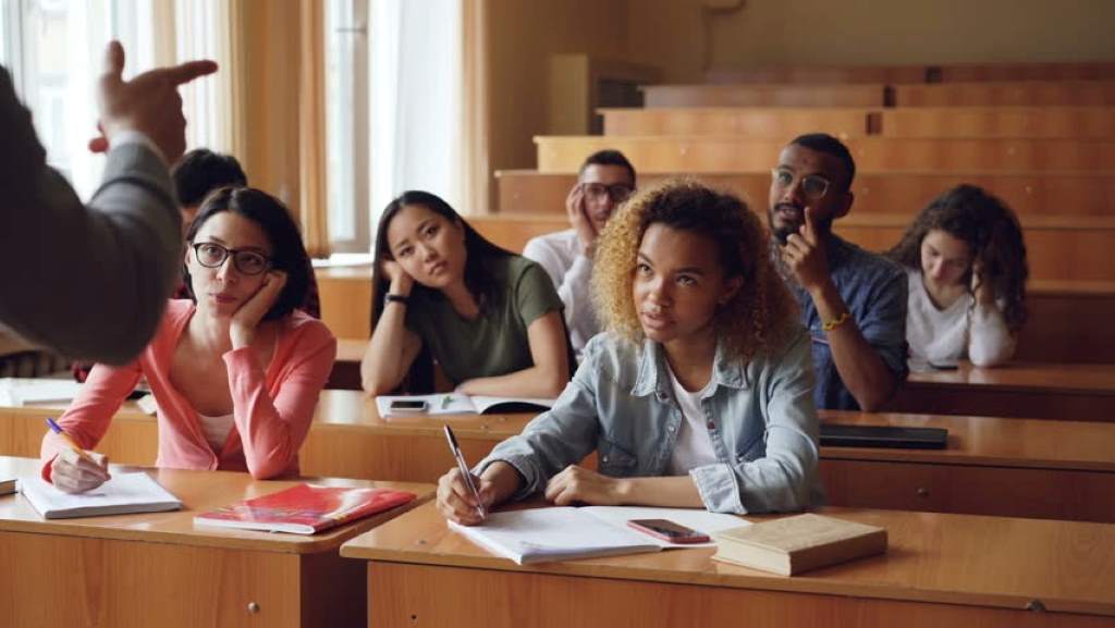 Teacher leading a standardized lecture while students appear disengaged and bored.
